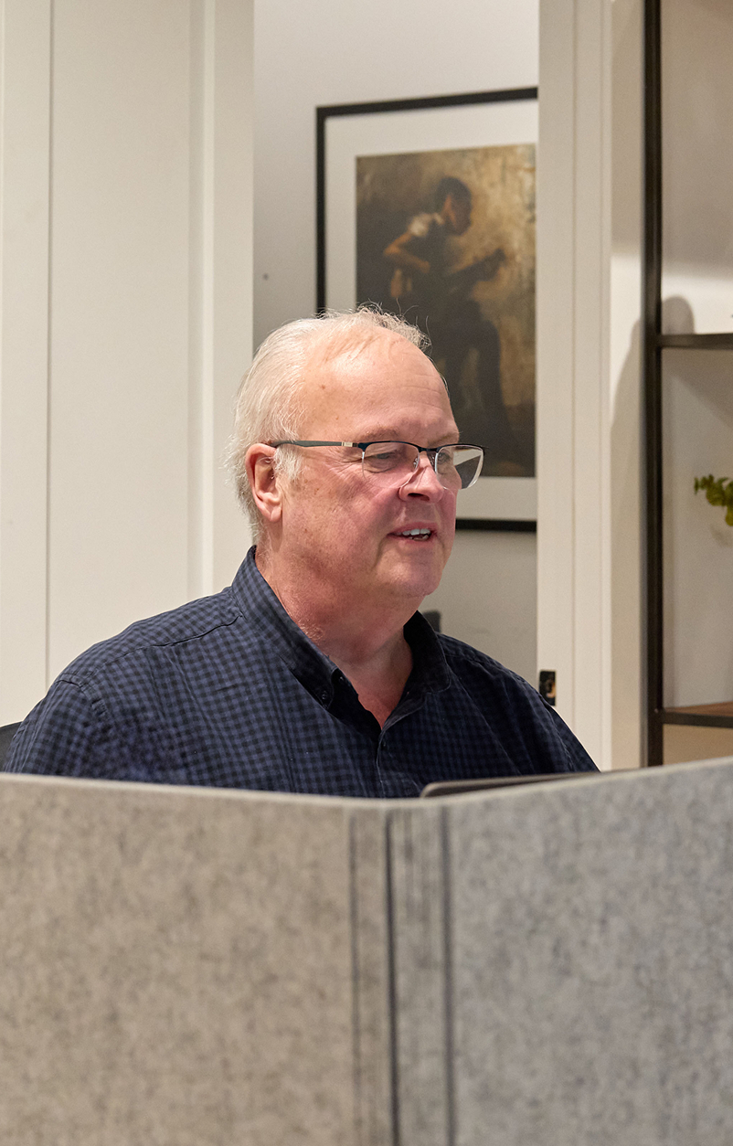 Older man with glasses and a checkered shirt sitting behind a gray partition in a room with framed art on the wall.
